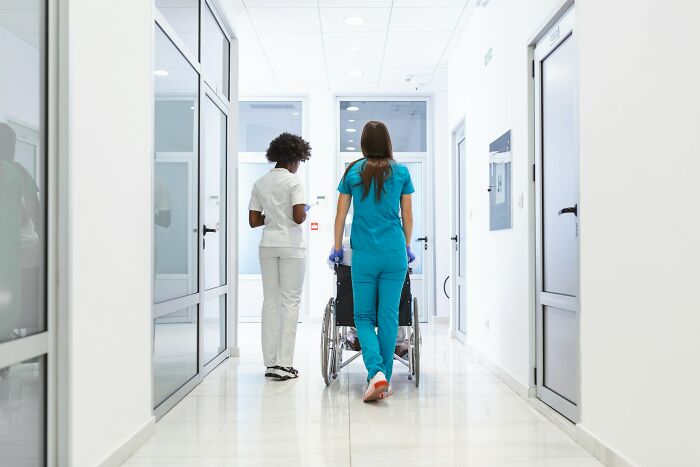 Two ER staff walking down a hospital hallway, one pushing an empty wheelchair, highlighting challenges in patient care.