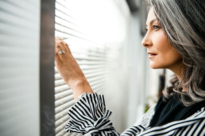 Middle-aged woman with gray hair peeking through window blinds, illustrating snobbish behavior shamed online.
