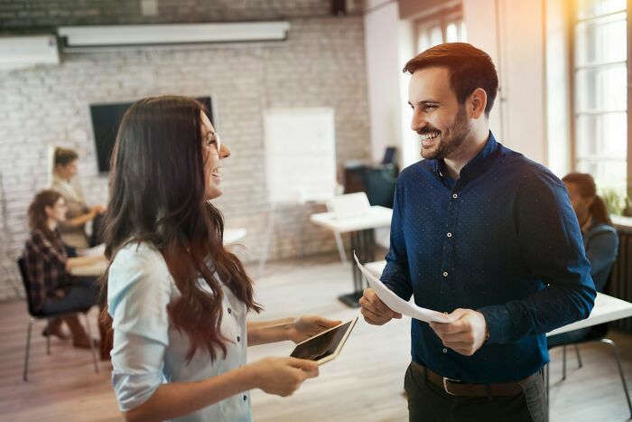 Two coworkers smiling and talking while holding documents and a tablet in an open office discussing workplace scandals.