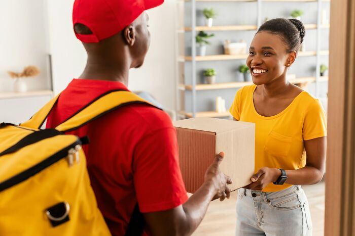 Postal worker in red delivering a package to a smiling woman at her door, highlighting realities of postal work.