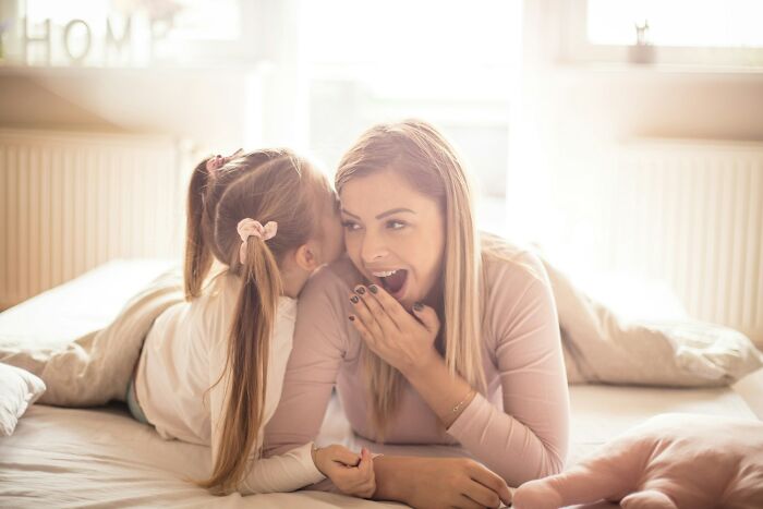 Mother and daughter sharing a secret in a bright bedroom, illustrating modern parenting trends and family bonding moments.