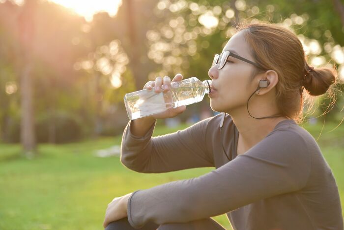 Young woman drinking water outdoors, wearing glasses and earbuds, enjoying a healthy lifestyle with surprising life facts.