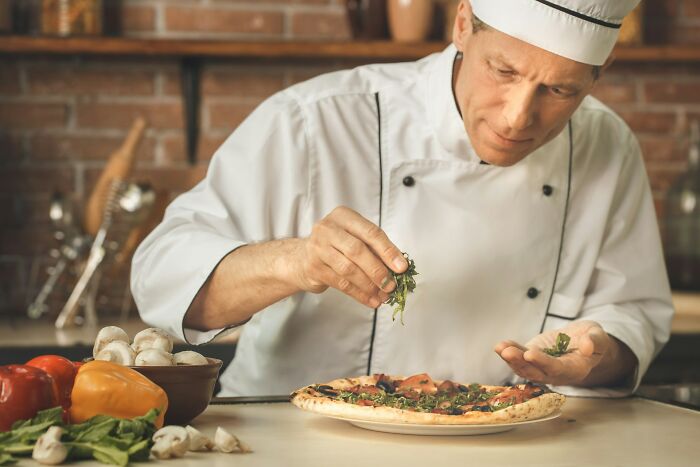 Chef in white uniform adding fresh herbs on a pizza, representing chefs who stopped cooking professionally moments.