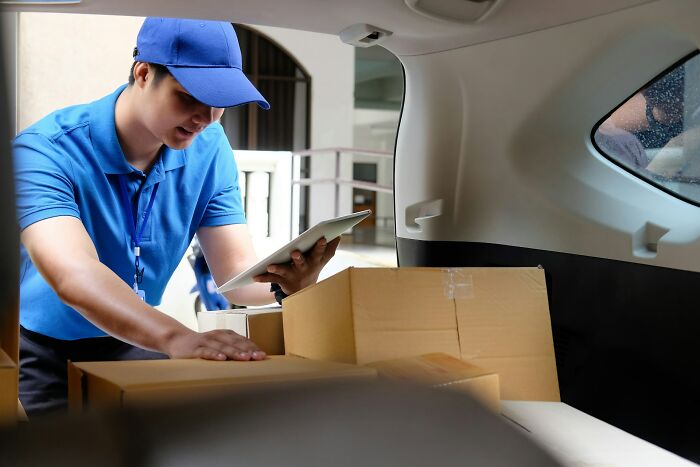 Postal worker in blue uniform checking deliveries on a tablet while loading packages into a vehicle trunk.