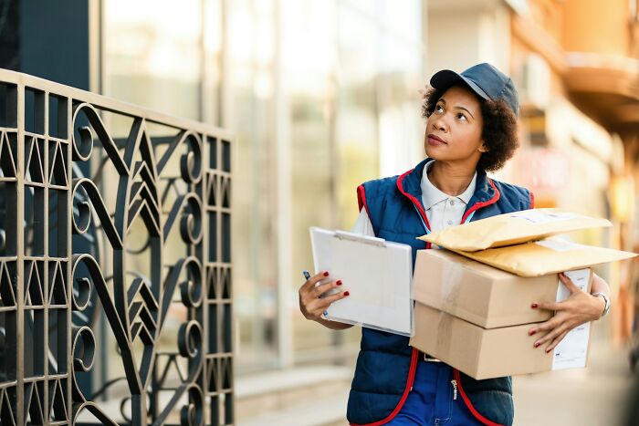 Postal worker carrying packages and clipboard while delivering mail on a city street during daytime.
