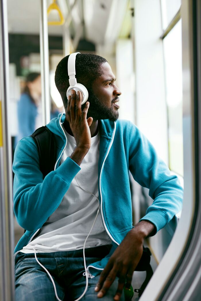 Man wearing white headphones and a blue hoodie, sitting by a window while enjoying a glitch in the system moment.