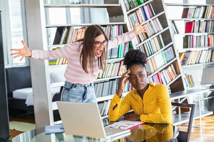 Frustrated employee wearing glasses angrily yelling at a coworker feeling stressed while working on a laptop in office.