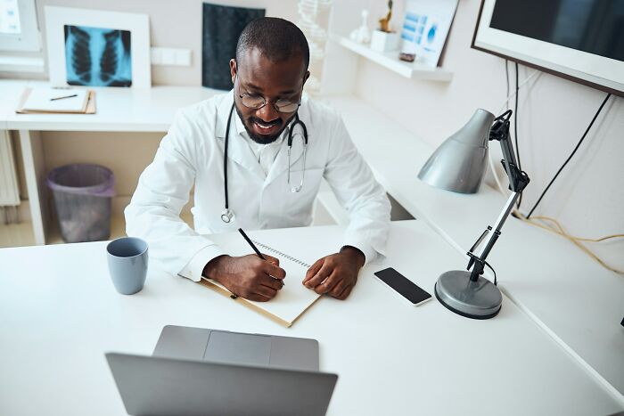 ER staff in white coat and glasses writing notes at desk with laptop and medical charts in bright office environment