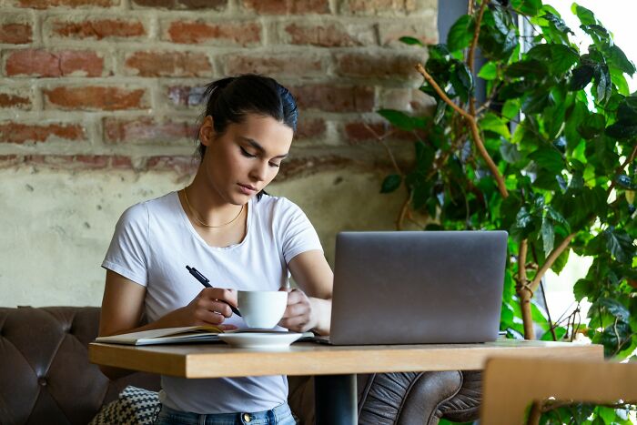 Young woman working on a laptop and taking notes in a cafe, showcasing the digital nomad lifestyle behind the scenes.