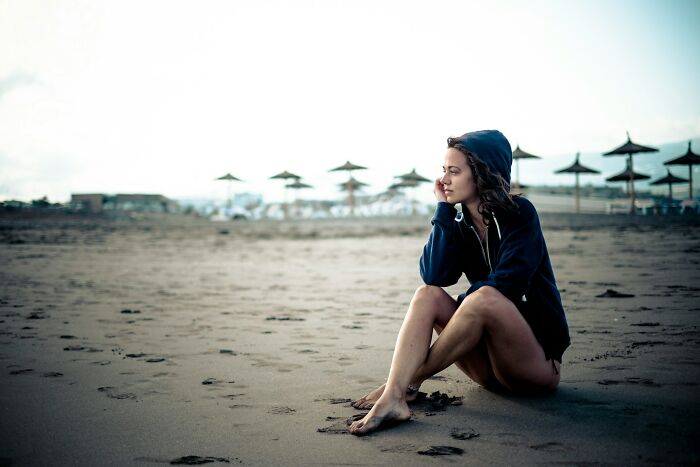 Young woman in a hoodie sitting alone on a deserted beach, capturing the hidden side of a digital nomad’s life.