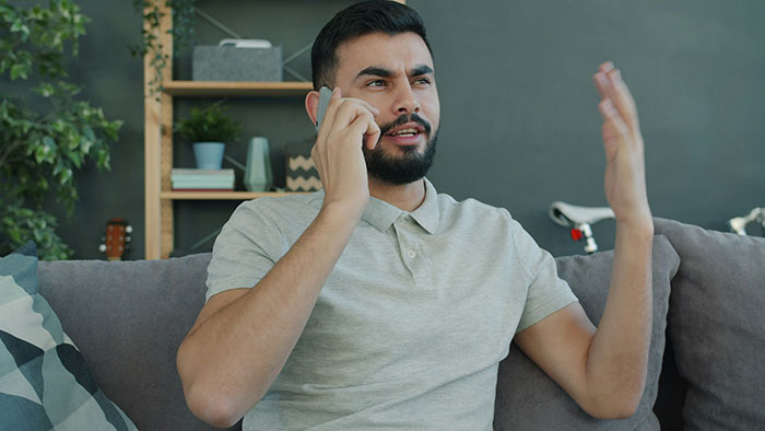 Man with beard sitting on couch, talking on phone with frustrated expression, illustrating gay bro treats sis like dirt.