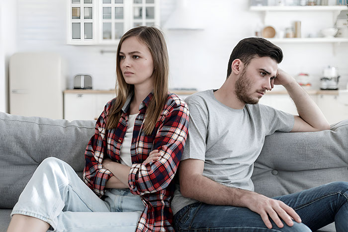 Upset couple sitting apart on couch after guy goes above and beyond for his ex girlfriend causing tension.