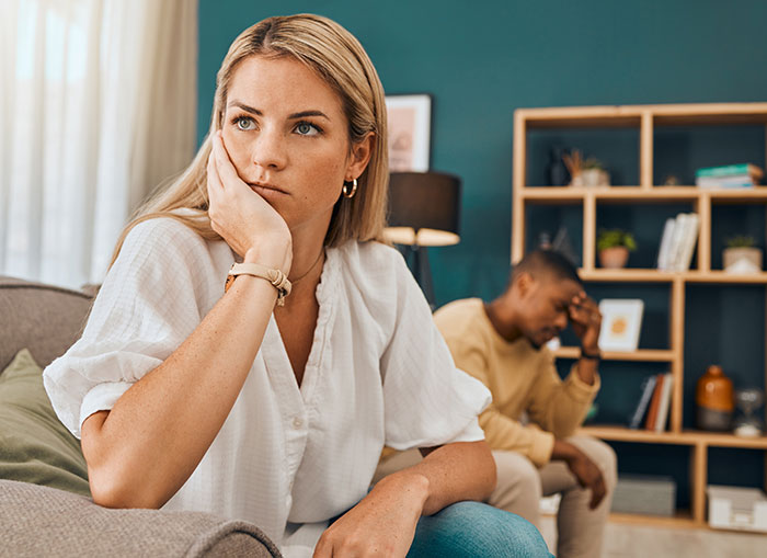 Upset woman looking away while man sits stressed in background, highlighting relationship tension and emotional conflict.
