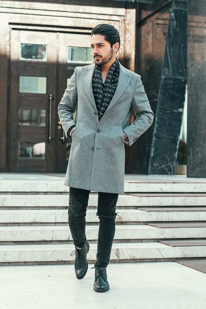 Young man dressed stylishly wearing a gray coat and black shoes, posing confidently outdoors on steps in the city.
