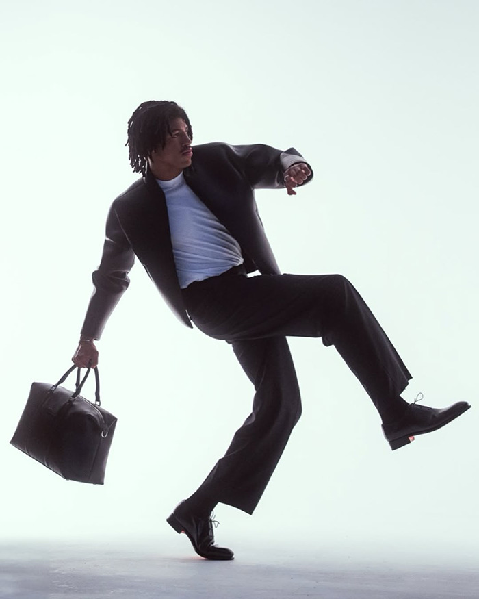 Young man dressed in black and white striking a dynamic pose with a large black bag, modeling fashion style.