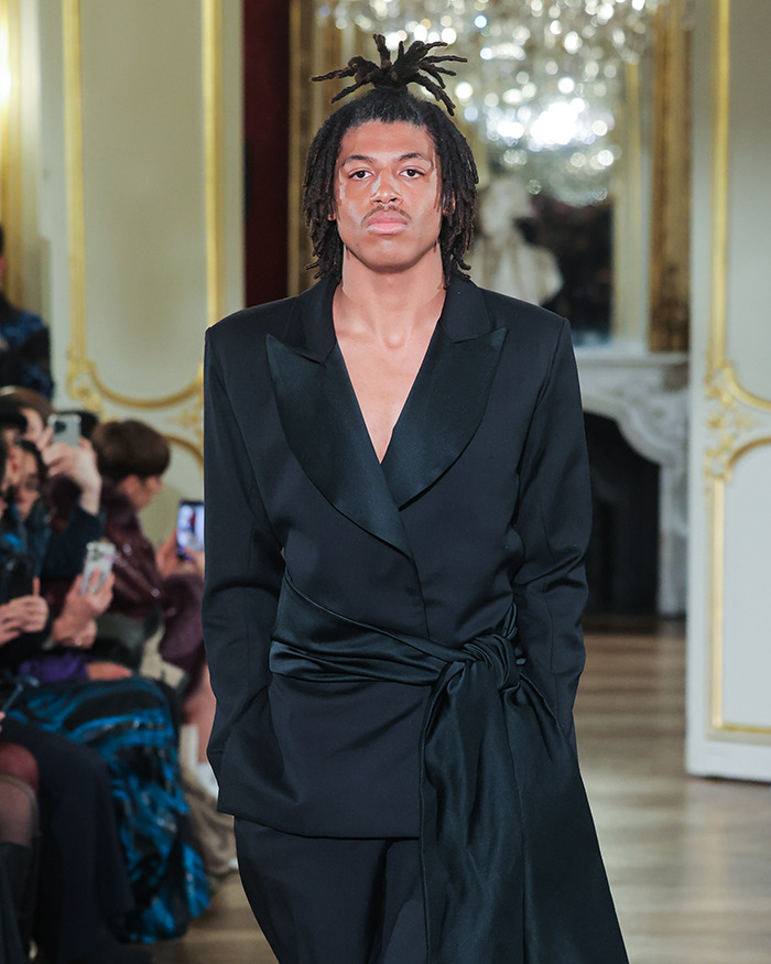 Young male model with dreadlocks wearing a black suit walking runway at a high-end fashion show event.