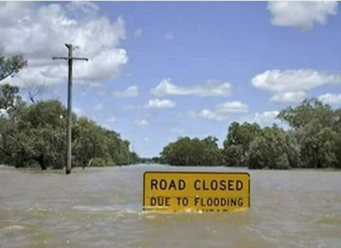 Flooded road with visible road closed sign surrounded by water under a partly cloudy sky, funny memes for dark cold winter days.