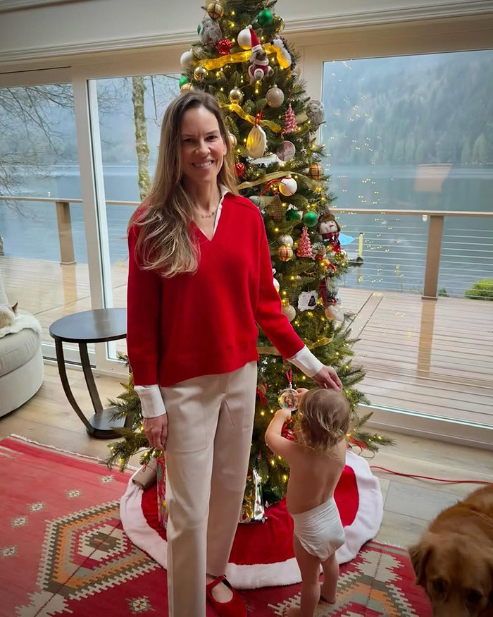 Woman in red sweater stands by Christmas tree with toddler decorating an ornament, relating to Hilary Swank and terminally ill kids.