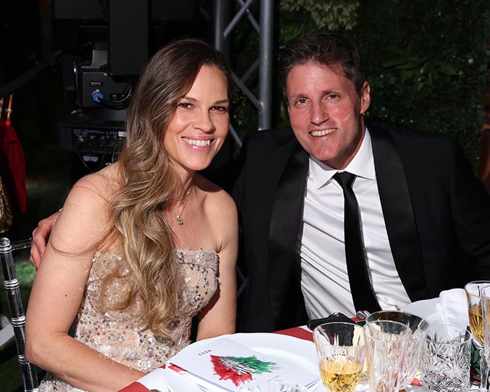 Hilary Swank smiles seated next to a man in a black suit at a formal dinner event outdoors.