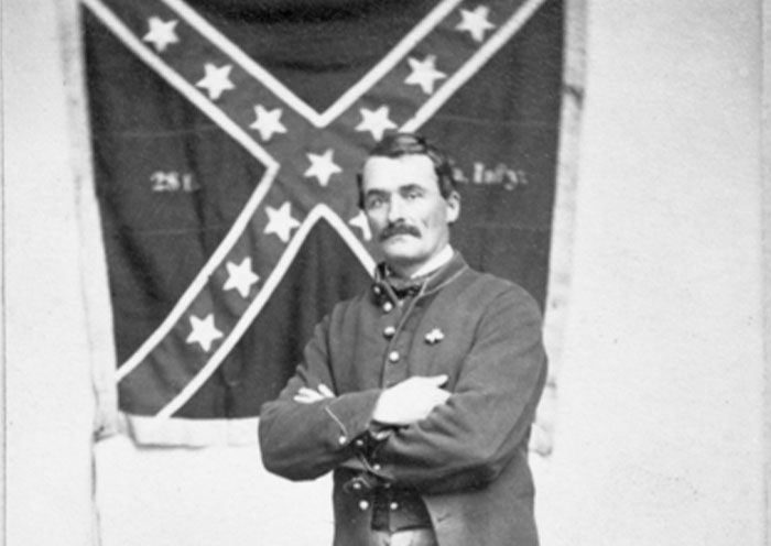 Black and white photo of a historical figure in uniform standing with arms crossed in front of a Confederate flag, bizarre history facts.
