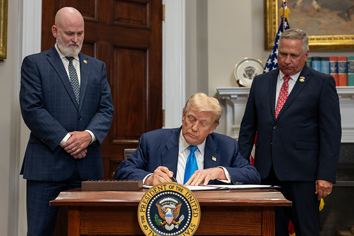 Former President signing documents in an official setting with two men standing nearby, symbolizing Epstein&rsquo;s private island home reveal.