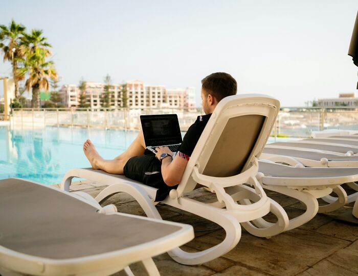 Man working on laptop by poolside on lounge chair, showcasing digital nomad life and remote work lifestyle outdoors.