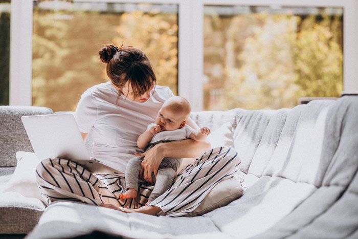 Stay-at-home mom holding baby on couch with laptop nearby, feeling overwhelmed by chores and parenting demands.