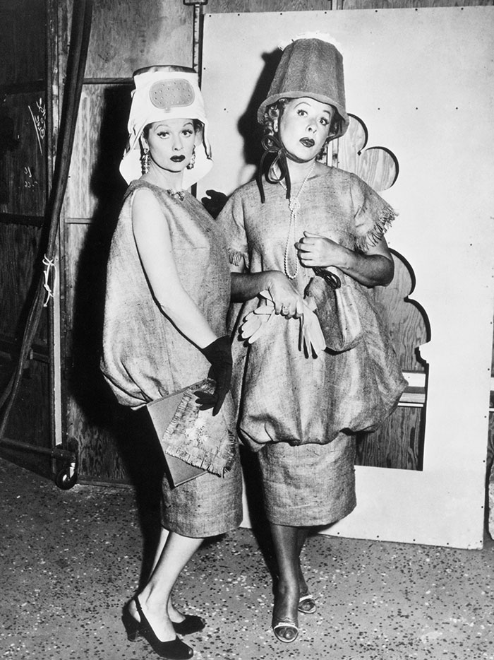 Two women in unusual hoop-style dresses and eccentric hats posing with gloves and handbags backstage.