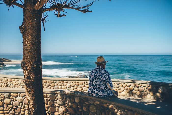 Man in a floral shirt and hat sitting by a stone wall, overlooking the ocean, reflecting on incredible minds without degrees.