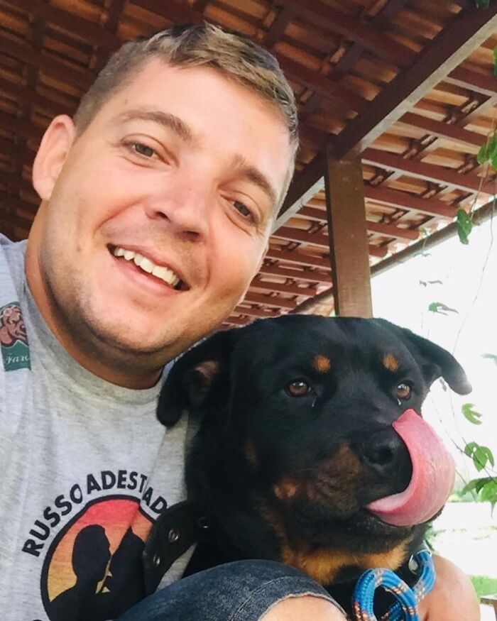 Smiling man taking a close selfie with a large black and brown dog wearing a blue collar under a wooden roof.