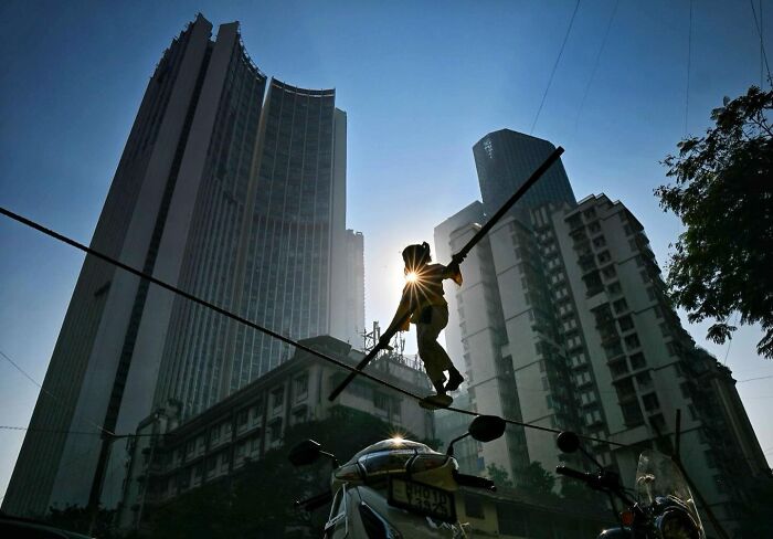 Tightrope walker balancing with poles between buildings in a bustling Mumbai street scene at sunrise.