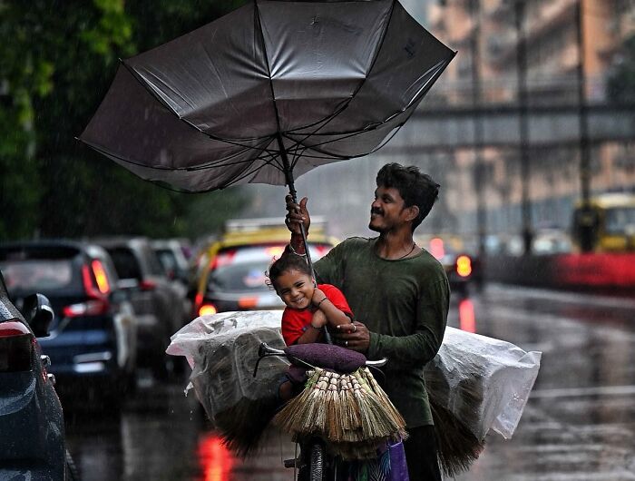 Man and child share umbrella on rainy Mumbai street with bicycles and traffic in the background capturing everyday life.