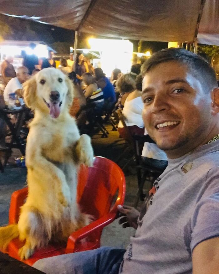 Man smiling and sitting next to a golden retriever dog in an outdoor dining area at night with people in the background