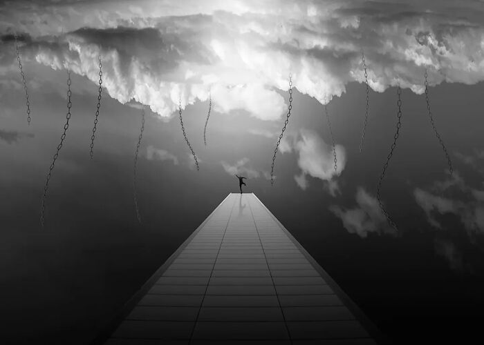 Minimalism art photo showing a person on a tall structure reaching toward sky with hanging chains in black and white.