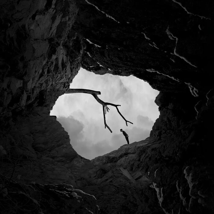 Minimalism art photograph showing a person standing near a barren tree branch inside a rocky cave opening under a cloudy sky.