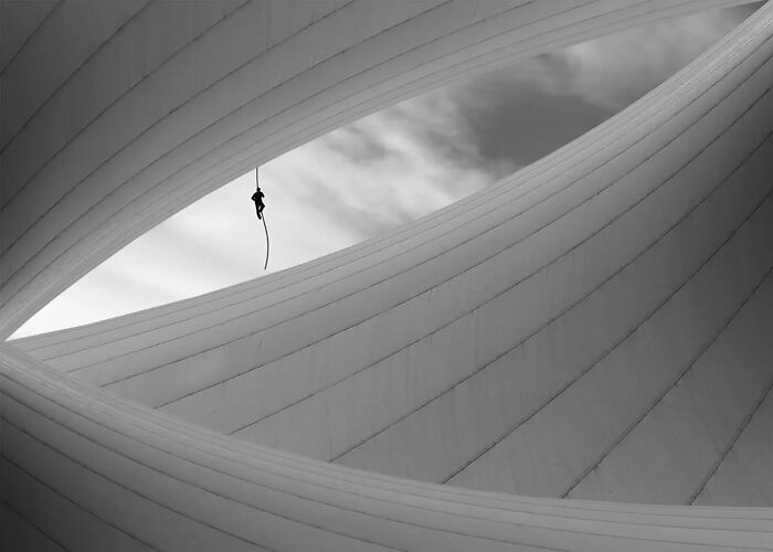 Minimalism art photo of a person hanging from a rope inside abstract curved architectural shapes in black and white.