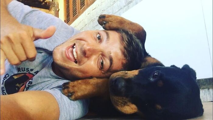 Man lying on the floor giving thumbs up while a dog rests its paw on his head showing close bond and affection.