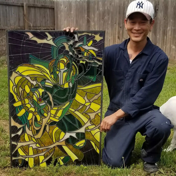 Artist posing outdoors with vivid stained glass sign depicting a colorful pop culture icon in yellow and green tones.