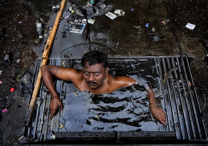 Man emerging from Mumbai street drain filled with water and trash, capturing everyday life in Mumbai street photos.
