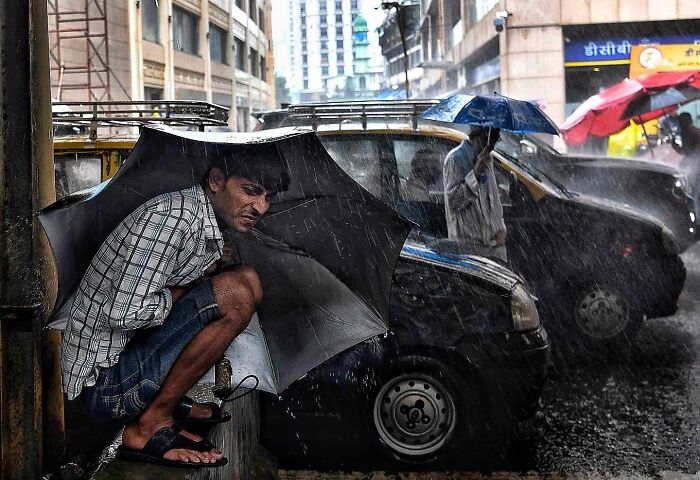 Man crouching under an umbrella on a rainy Mumbai street, capturing everyday life in vibrant urban scenes.