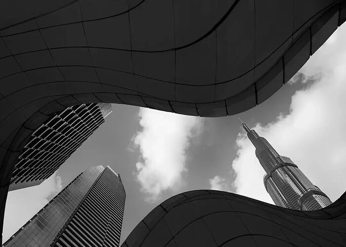 Black and white minimalist photo showing modern skyscrapers framed by curving architectural shapes against a cloudy sky.