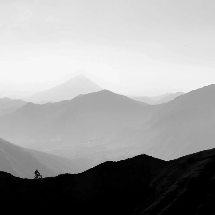 Minimalism photography showing a lone cyclist on a mountain ridge with layered hills and a distant peak in soft grayscale.