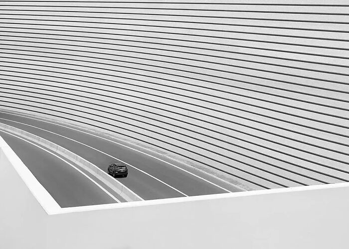 Minimalism in photography shown by a lone car driving on a curved road against a striped architectural backdrop.