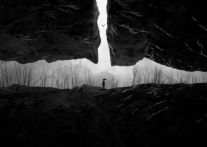 Black and white minimalist photograph of a person holding an umbrella between large rock formations under a flying bird.