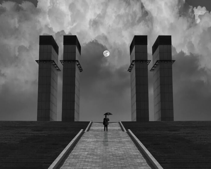 Minimalist black and white photo of a person with umbrella walking towards tall pillars under a cloudy sky and full moon.