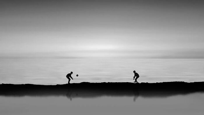 Two silhouetted figures playing ball on a narrow strip of land with a minimalist seascape background in black and white.