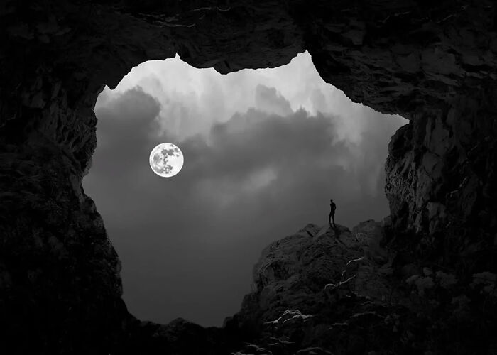 Black and white minimalist photograph of a person standing on rock inside a cave with a full moon in the sky.