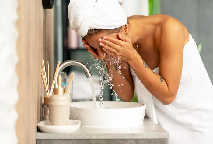 Woman washing face at sink wrapped in towel showing human body features and daily self-care routine.