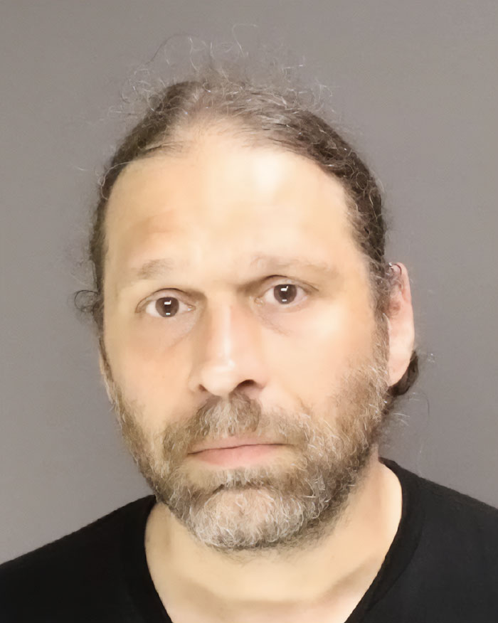 Man with long hair and beard, wearing a black shirt, shown in a close-up mugshot related to NYU shove victim viral video.