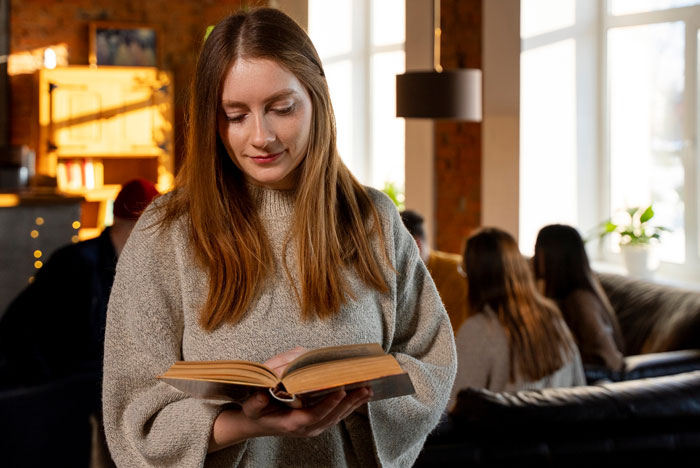 Young woman reading a book indoors with a group in background, related to Jehovah's Witness mom celebrating kid's birthday under new name.